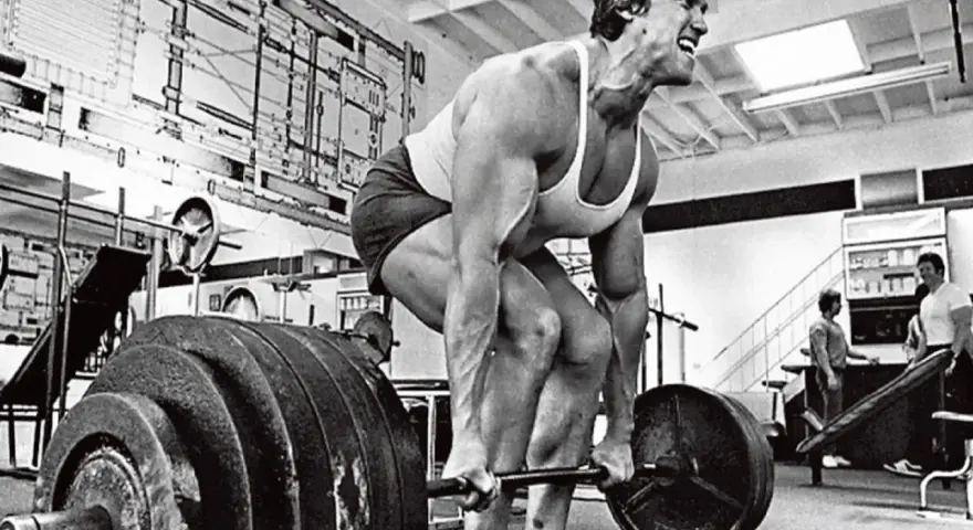 Arnold Schwarzenegger esegue uno stiff legged deadlift in palestra, concentrato sul sollevamento del bilanciere.