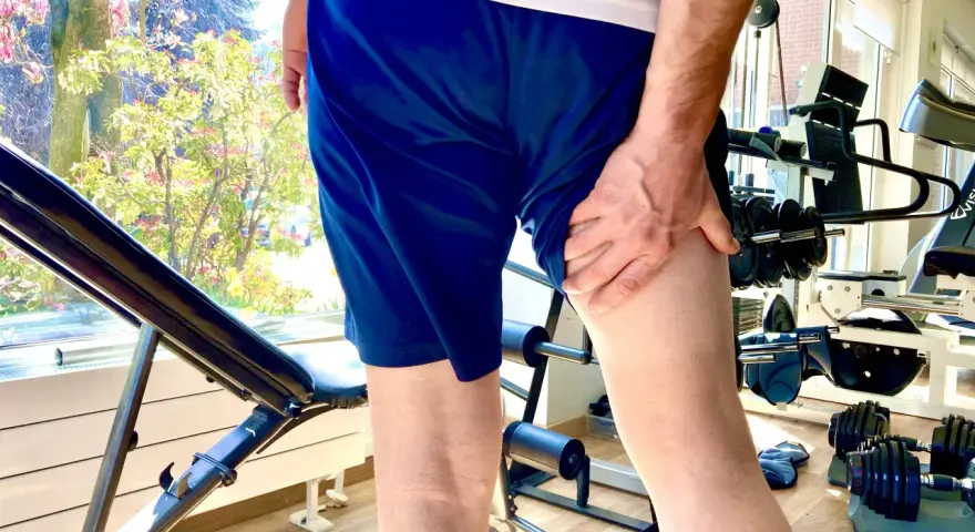 Uomo con mano sulla coscia, indicando una possibile lesione hamstring. Attrezzatura da palestra e alberi fioriti sullo sfondo.