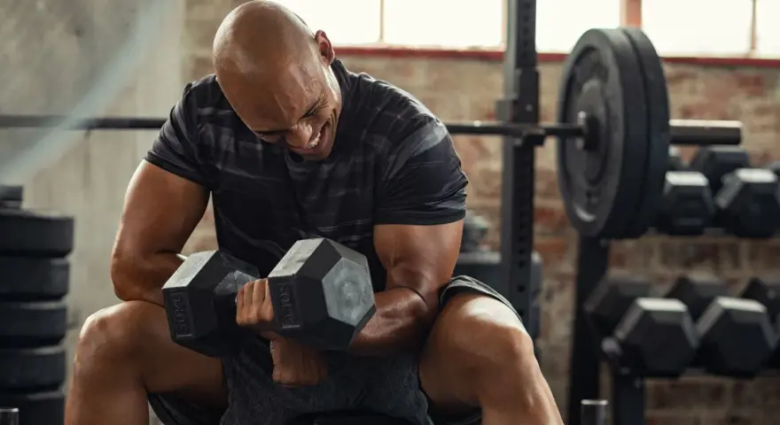Uomo calvo in palestra, concentrato su un curl con manubri da 50 kg. Il suo viso mostra lo sforzo, indicando un RPE chart elevato.