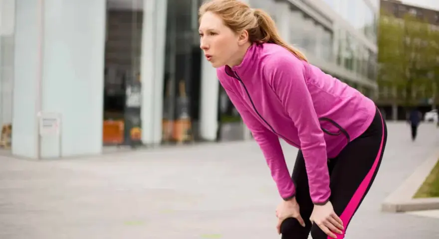 Donna bionda, esausta dopo la corsa, si piega appoggiando le mani sulle ginocchia. Il suo respiro affannoso indica un alto RPE (Rate of Perceived Exertion).