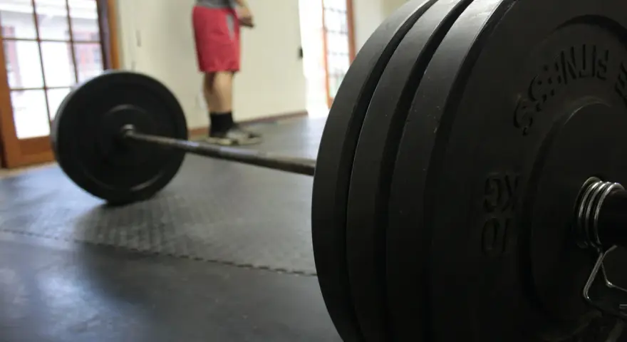 Bilanciere con pesi pronti per un allenamento sulla panca piana. Un uomo in pantaloncini rossi attende sullo sfondo.
