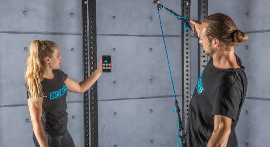 Donna con smartphone e uomo con cavo per allenamento. Il display mostra grafici, forse per misurare la forza o il progresso, spiegando cosa significa pr.