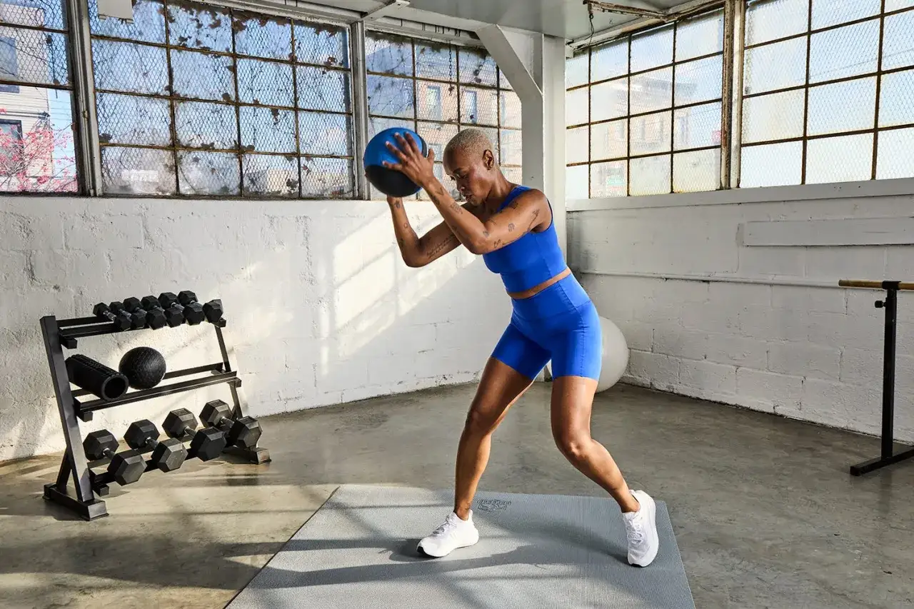 Donna esegue esercizi con la palla medica in palestra, con pesi e foam roller sullo sfondo.