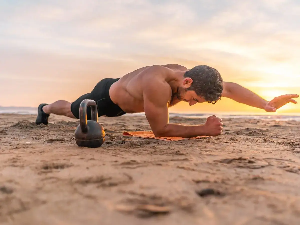 Uomo in posizione di plank sulla spiaggia al tramonto, con un kettlebell accanto. Un allenamento efficace per 10 esercizi addominali.