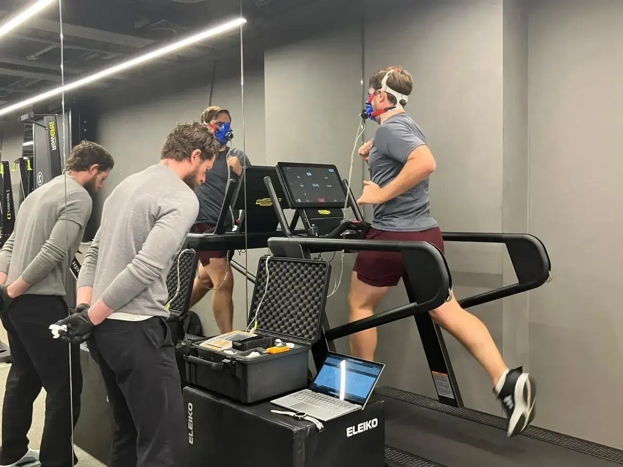 Atleta corre su tapis roulant con maschera per misurare i vo2 max valori. Due uomini osservano i dati su un laptop.