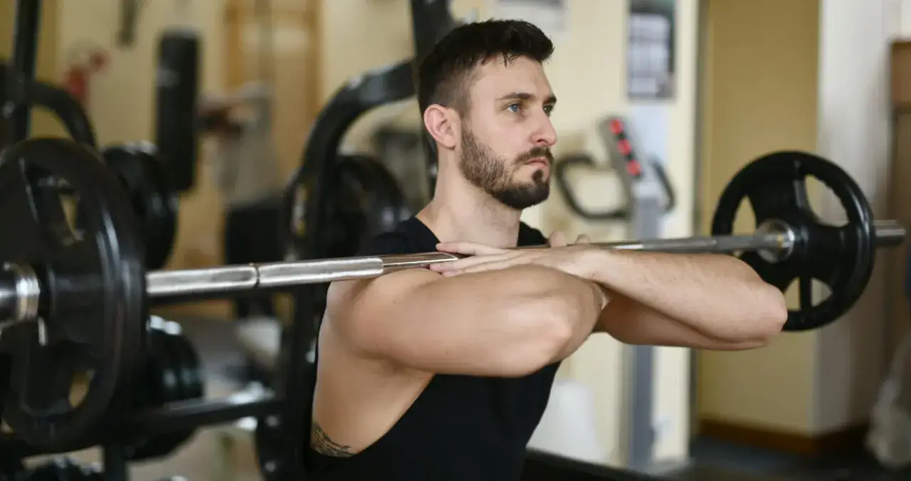 Uomo con barba esegue un front squat multipower in palestra, con bilanciere appoggiato sulle spalle.