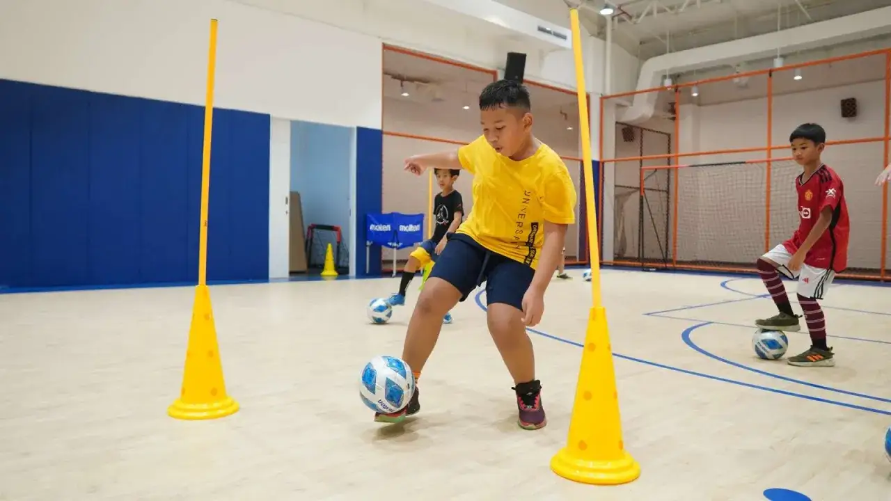 Bambini si allenano in un circuito forza calcio, dribblando coni gialli e palloni.