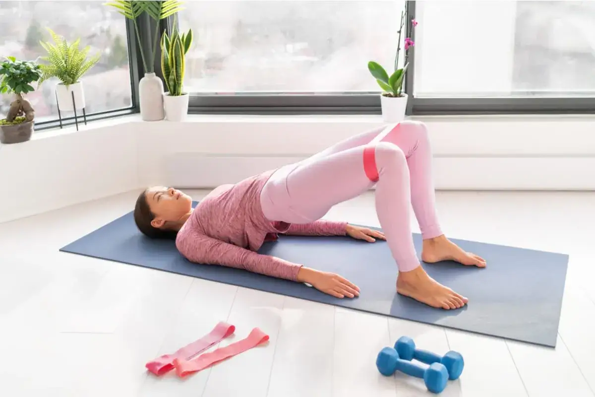 Donna esegue hip thrust a casa con elastico rosa sulle cosce. Sul tappetino, vicino a due manubri blu, si allena per glutei forti.