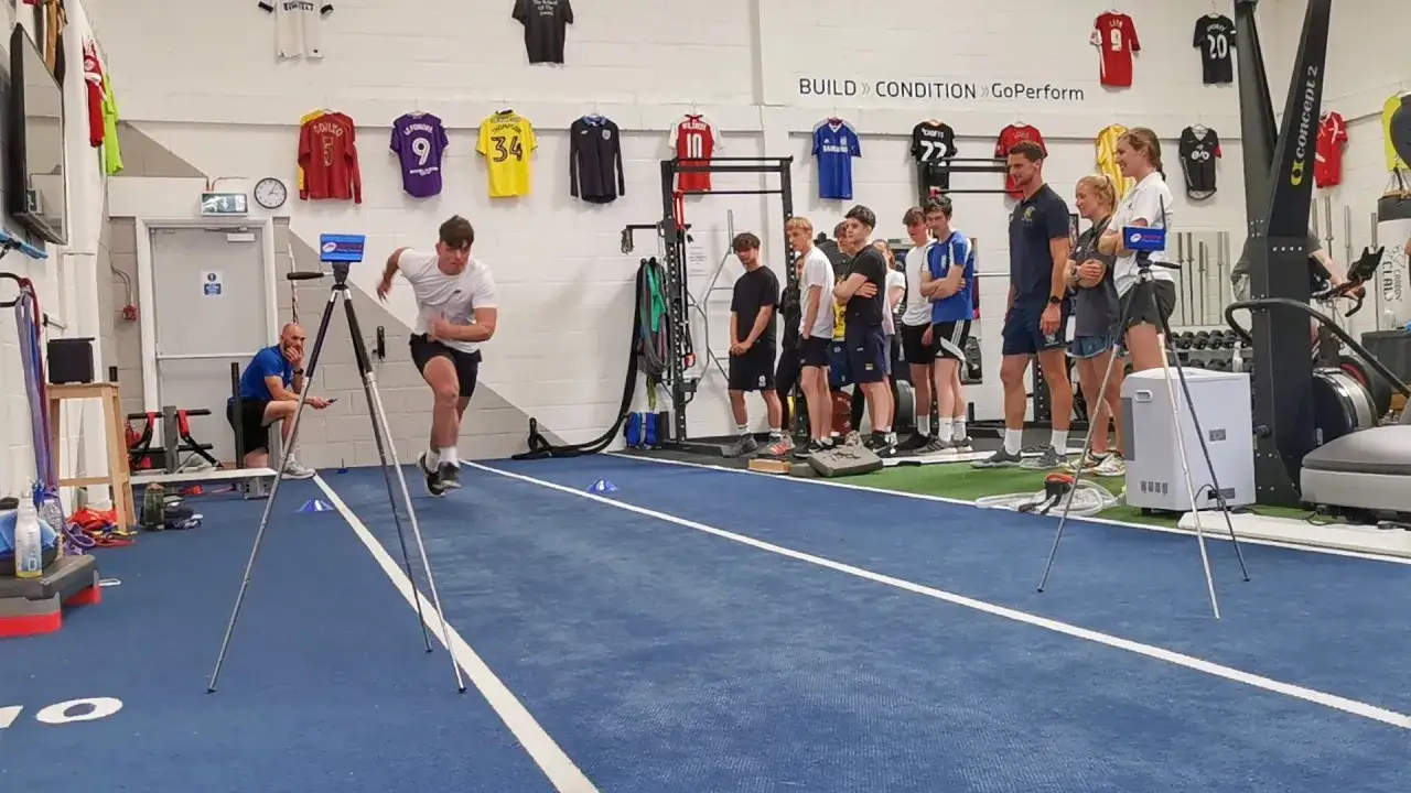Giovane atleta esegue uno sprint durante un fitness test, osservato da allenatori e compagni in una palestra attrezzata.
