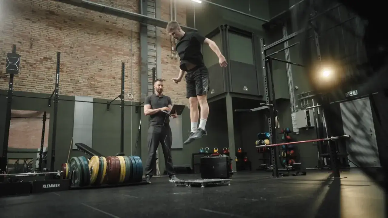 Atleta in aria durante un countermovement jump, mentre un allenatore osserva i dati su un laptop.