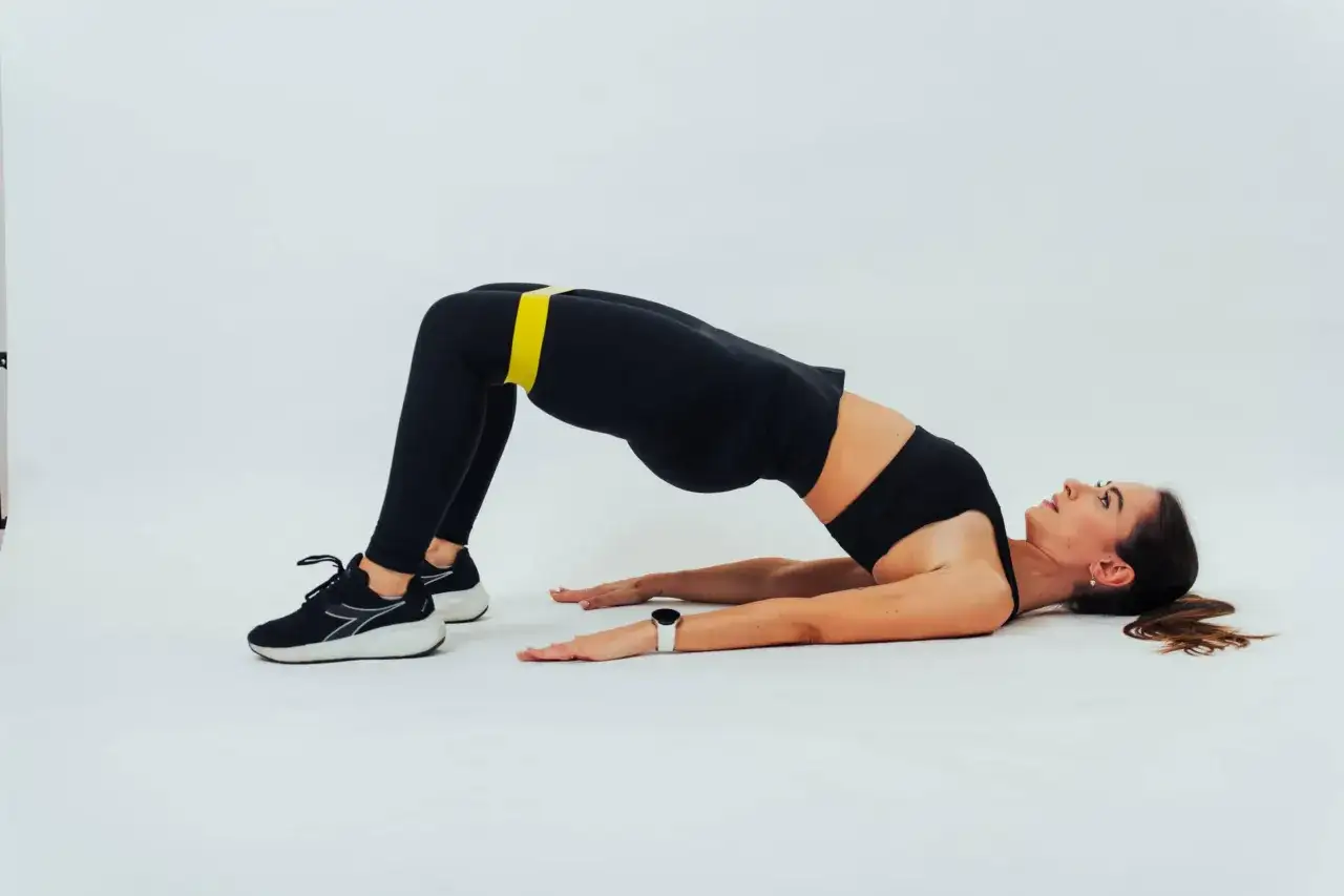Donna esegue un hamstring bridge con elastico giallo sulle cosce. Schiena inarcata, braccia distese a terra.