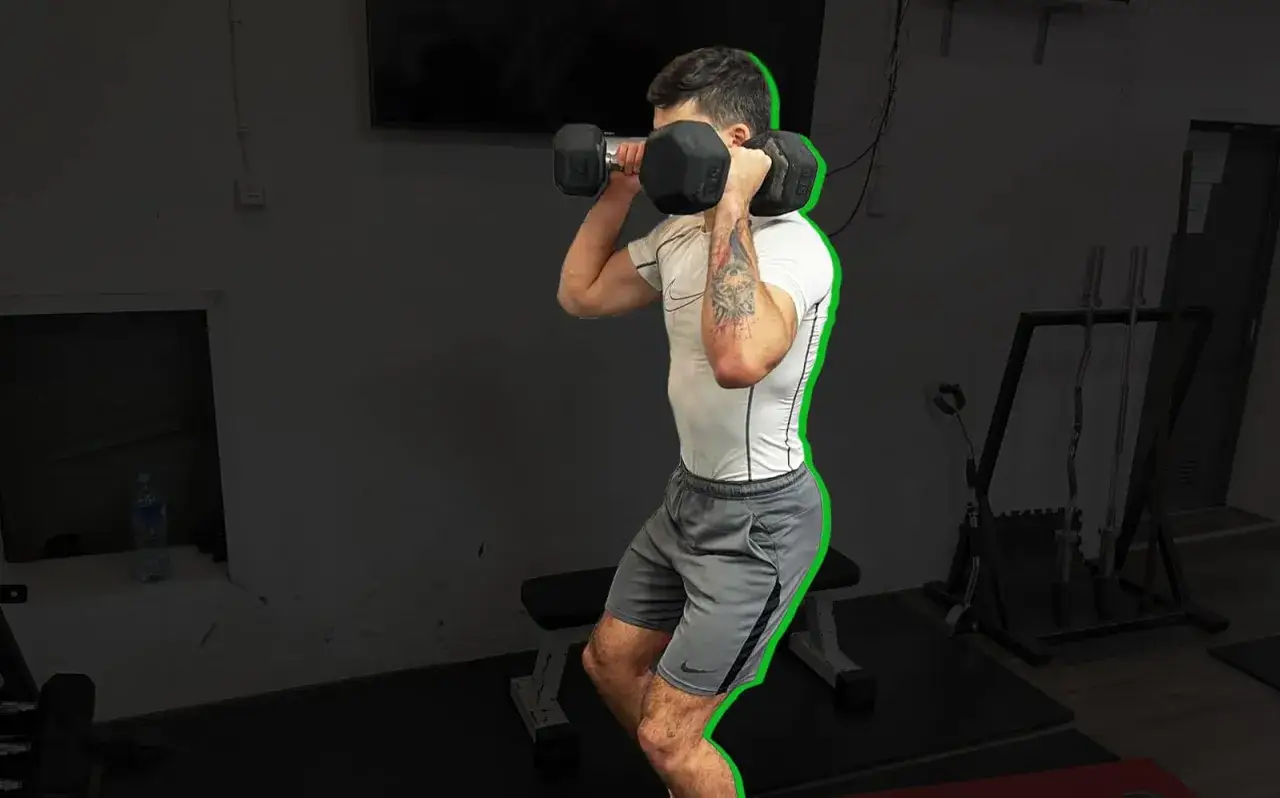 Uomo in palestra esegue push press con manubri, con le braccia piegate e i pesi all'altezza delle spalle, pronto a spingere.